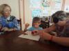 The boy is sitting  at the table. On the table is a braille board. Two women beside him. One with a blindfold